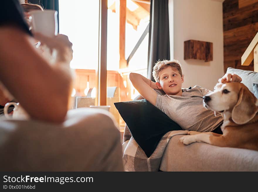 Cozy family tea time. Father and son at the home living room. Boy lying on comfortable sofa and  stroking their beagle dog and smiling. Peaceful family moments concept image