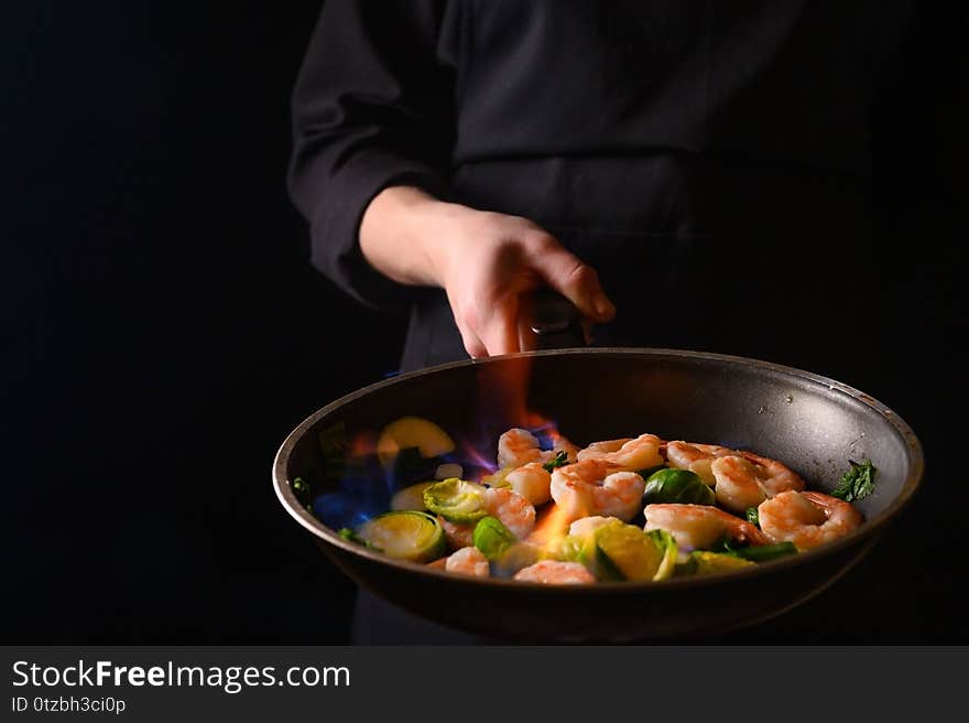 Professional chef cook on a dark background prepares seafood, shrimp with fire and vegetables. Roasting and cooking veggie food. Black background, Space for design.