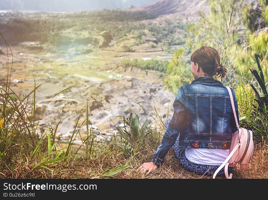 Young woman sitting on a rock with backpack and looking to the horizon. Bali island. Volcano Batur. Indonesia.