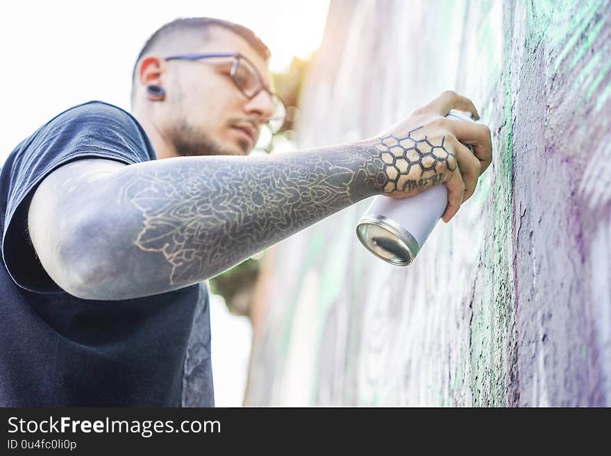 Tattooed graffiti artist painting with color aerosol on the wall - Contemporary spray write at work - Urban lifestyle,street art concept - Focus on his hand can