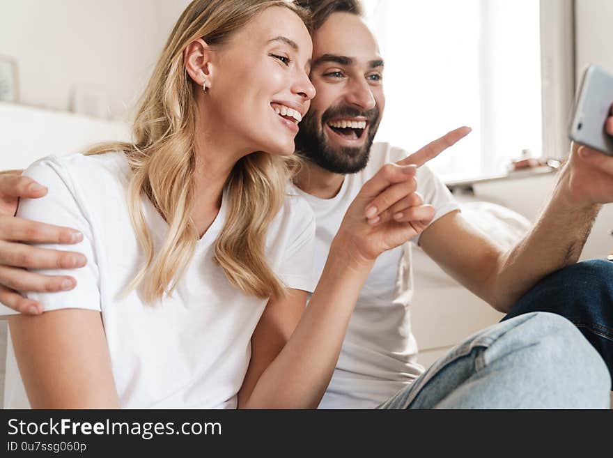 Cheerful beautiful young couple sitting at the couch on a floor at home, holding mobile phone