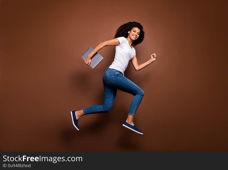 Full length profile photo of funny dark skin lady jumping high holding, notebook hurry classes lessons schoolgirl wear casual white t-shirt jeans isolated brown color background. Full length profile photo of funny dark skin lady jumping high holding, notebook hurry classes lessons schoolgirl wear casual white t-shirt jeans isolated brown color background