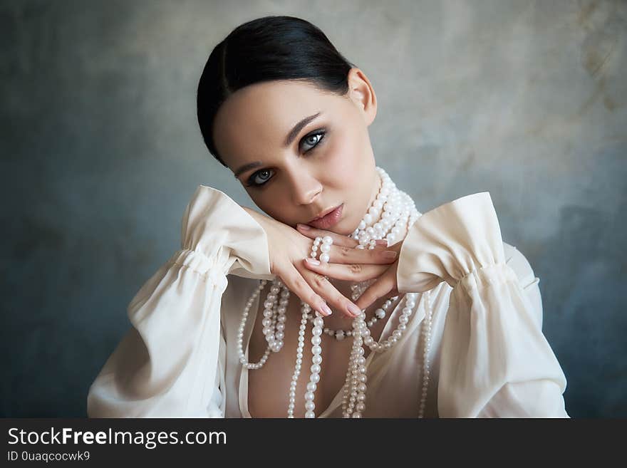 Girl dressed in white boho clothing with white pearl beads around her neck is sitting at a table. Perfect smile, romantic sexy image of a woman, clean smooth skin and beautiful makeup