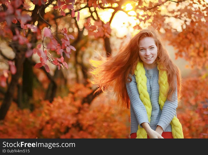 A woman stands in a forest with falling red leaves and looks directly at the camera.