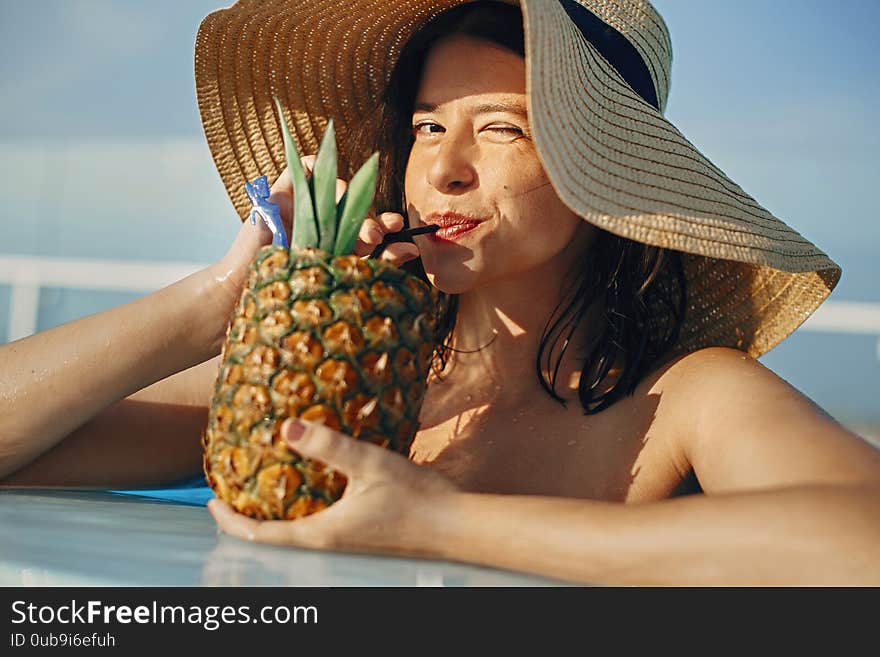 Beautiful young woman smiling and drinking cocktail in pineapple, relaxing in pool on summer vacation. Girl in hat enjoying warm sunshine with drink in pool on rooftop in luxury tropical resort