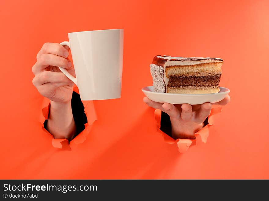 Hand holding a mug of coffee and a piece of chocolate cake through torn holes in red paper background.