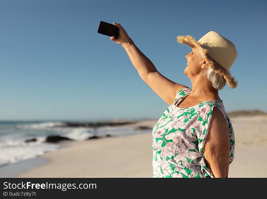 Side view of a senior Caucasian woman at the beach in the sun, wearing a sun hat, holding a smartphone, taking a selfie and smiling, with sea and blue sky in the background. Side view of a senior Caucasian woman at the beach in the sun, wearing a sun hat, holding a smartphone, taking a selfie and smiling, with sea and blue sky in the background