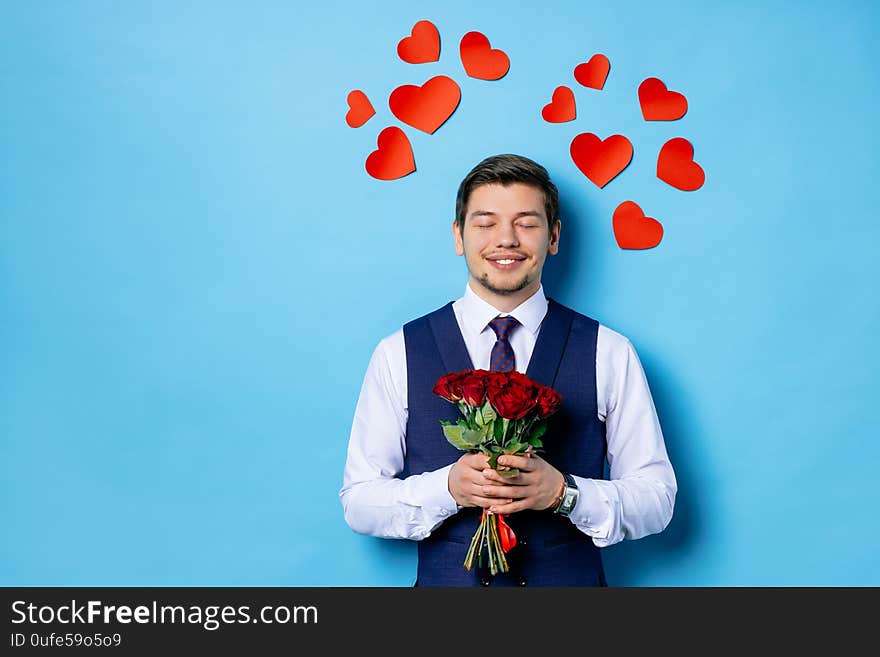 Guy in tuxedo isolated over blue background. smiling caucasian male in formal wear stand with closed eyes holding red roses in hands and dream, waiting for valentines day. love, celebration concept. Guy in tuxedo isolated over blue background. smiling caucasian male in formal wear stand with closed eyes holding red roses in hands and dream, waiting for valentines day. love, celebration concept