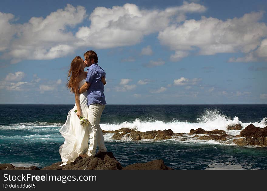 Young just married couple kissing on ocean rock shore. Side view. Ocean waves, splash of water and blue cloudy sky. Wedding ceremony on ocean coast