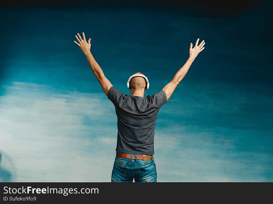 A bald man in headphones listens to music, standing with his back to the camera, raising his hands up on a blue