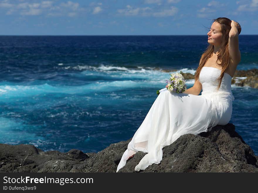 Young elegant bride in light white wedding dress sitting on ocean rock shore, flowers in hand. Ocean waves, splash of water and blue cloudy sky. Wedding ceremony on ocean coast. Young elegant bride in light white wedding dress sitting on ocean rock shore, flowers in hand. Ocean waves, splash of water and blue cloudy sky. Wedding ceremony on ocean coast