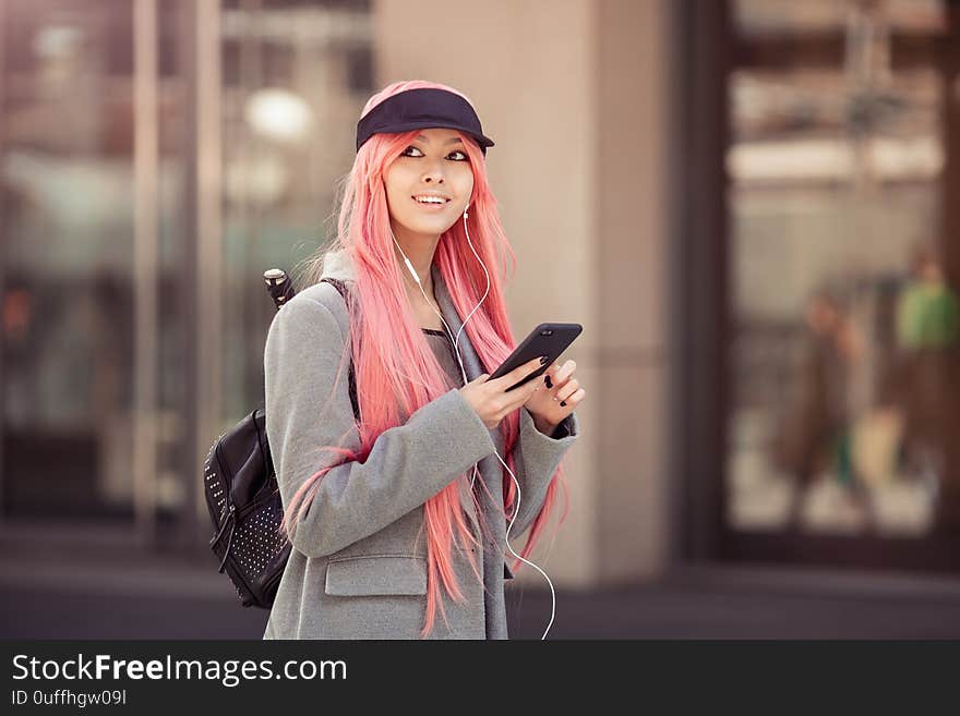 Japan anime cosplay. Colorful portrait of a young attractive smiling Asian woman in a coat with creative make-up wearing a pink wig. Trendy Japanese girl using smartphone listening music in earphones walking on the city street. Japan anime cosplay. Colorful portrait of a young attractive smiling Asian woman in a coat with creative make-up wearing a pink wig. Trendy Japanese girl using smartphone listening music in earphones walking on the city street.