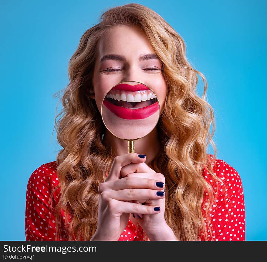 Blonde holds a magnifying glass in front of her teeth. Girl with a bright smile. Funny girl. Young woman holds a magnifying glass in front of her teeth. More teeth. Dental care. Funny face of a girl