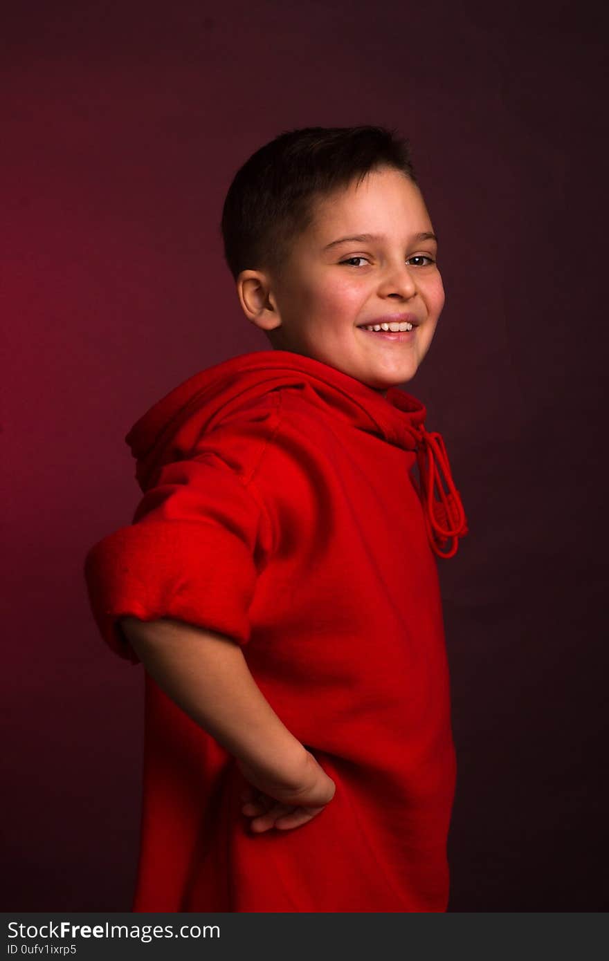 Studio portrait of a dark-haired boy, on a maroon background, in a red hoodie jacket. Positive emotion, smile on your face