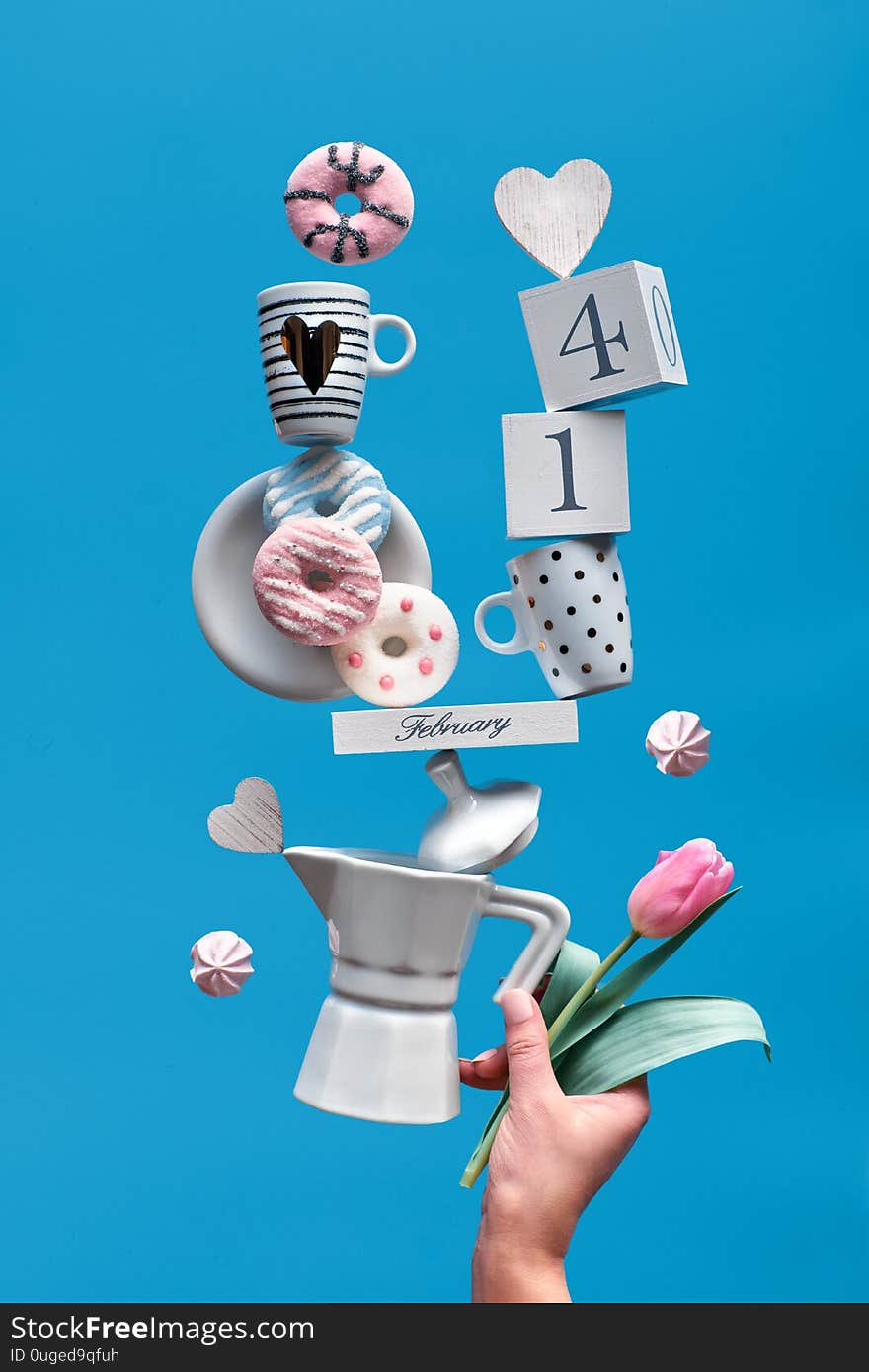 St. Valentine`s day fun background in blue mint . Date 14 February on wooden calendar with coffee cups, tulip, pink donuts and marshmallows. Female hand holding balancing pyramid on coffee maker