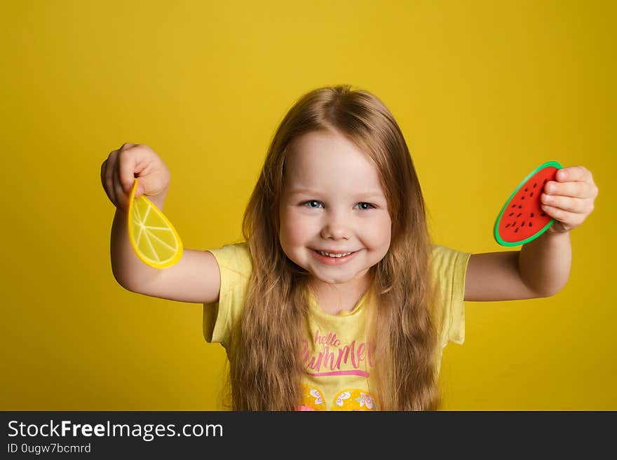 Front view of happy girl standing on yellow isolated background and closing eyes with toys. Cheerful longhaired child laughing and playing hide and seek. Concept of game and happiness. Front view of happy girl standing on yellow isolated background and closing eyes with toys. Cheerful longhaired child laughing and playing hide and seek. Concept of game and happiness.