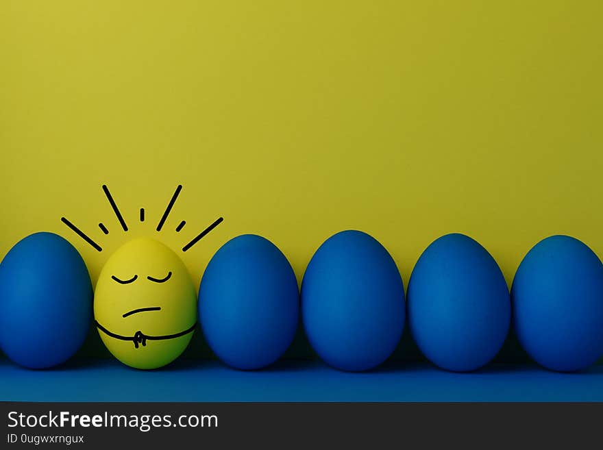 Five blue and one yellow funny faced painted Easter eggs stand in a row on a yellow with blue background.