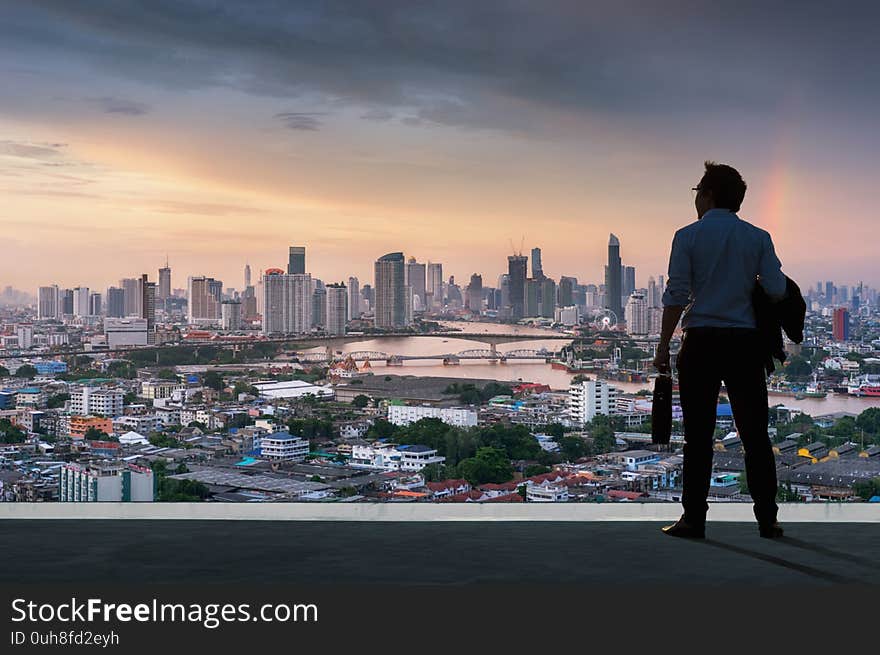 Concept vision, Young businessman wearing comfortable casual suit jacket standing holding a work bag on top of building and looking through the modern buildings in the skyline. Concept vision, Young businessman wearing comfortable casual suit jacket standing holding a work bag on top of building and looking through the modern buildings in the skyline