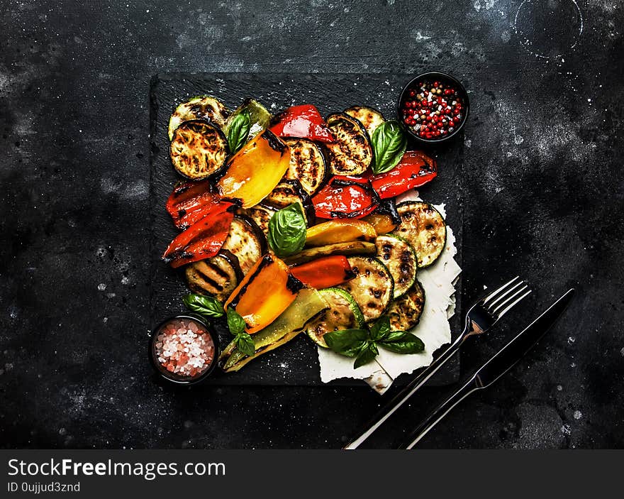 Grilled multicolored vegetables, aubergines, zucchini, pepper with green basil on serving stone board on black background, top