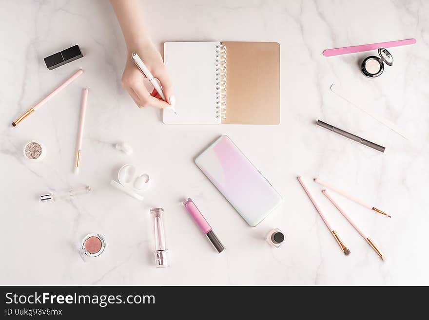 Feminine workspace in flat lay style with female hand writing in notepad, up-to-date smartphone mockup, cosmetics and manicure products on marble background. Woman`s freelance concept. Copy space. Mockup for your design.