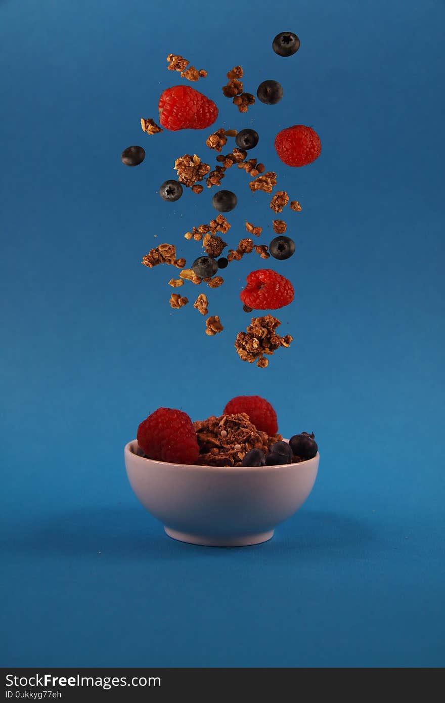 Granola and berries pouring to the white bowl against blue background. Image contains copy space. Concept of flying food