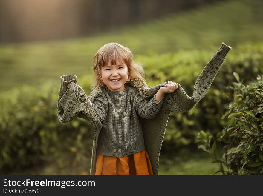 Baby Girl In Warm Clothes In The Field With Tea Tree Plants.