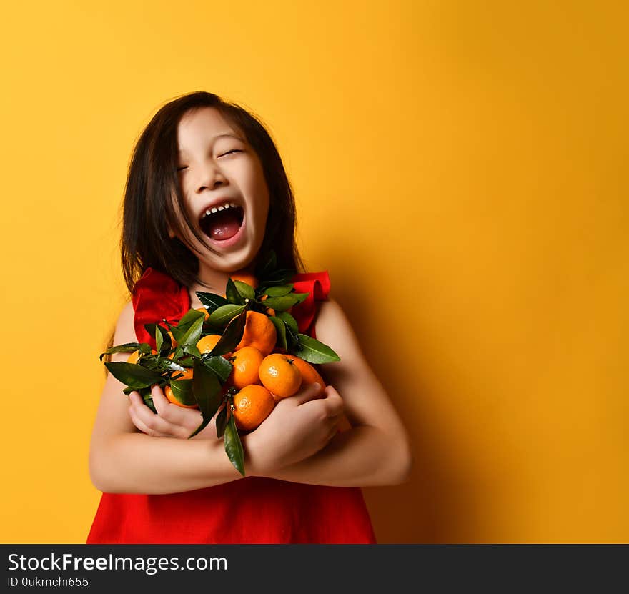 Asian kid in red blouse. Laughing out loud, holding an armful of tangerines and oranges, posing on orange background. Close up