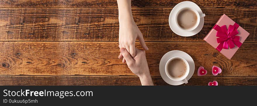 Valentine`s Day celebration concept. A nice gift for your loved one. Hands of man and woman with coffee mugs on a wooden table background. Copy space. Flat lay