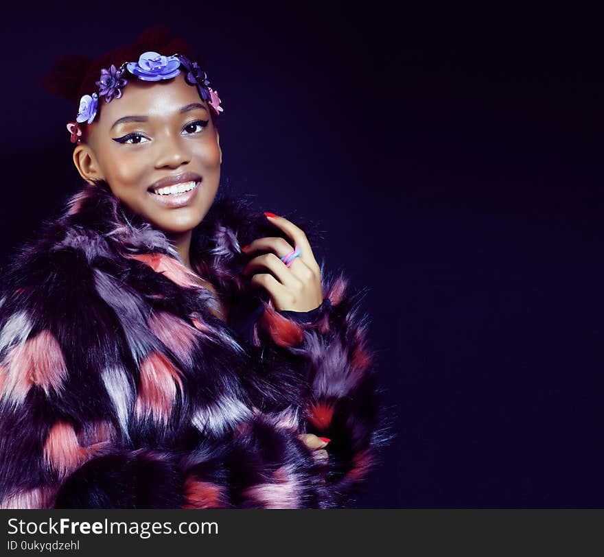 Young pretty african american woman in spotted fur coat and flowers on head smiling on black background, fashion people concept closeup