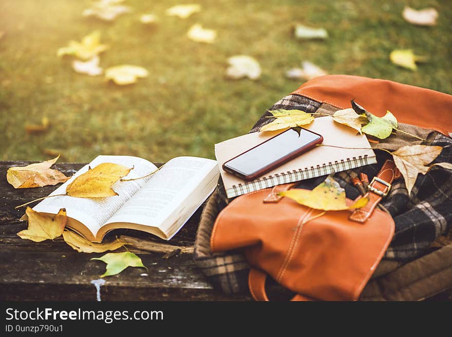 The backpack is placed on an antique wooden table with books, notebooks, phones and the nature of maple leaves from the top view