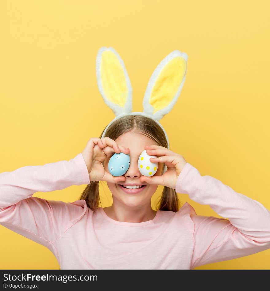 Cheerful child with bunny ears covering eyes with easter eggs isolated on yellow