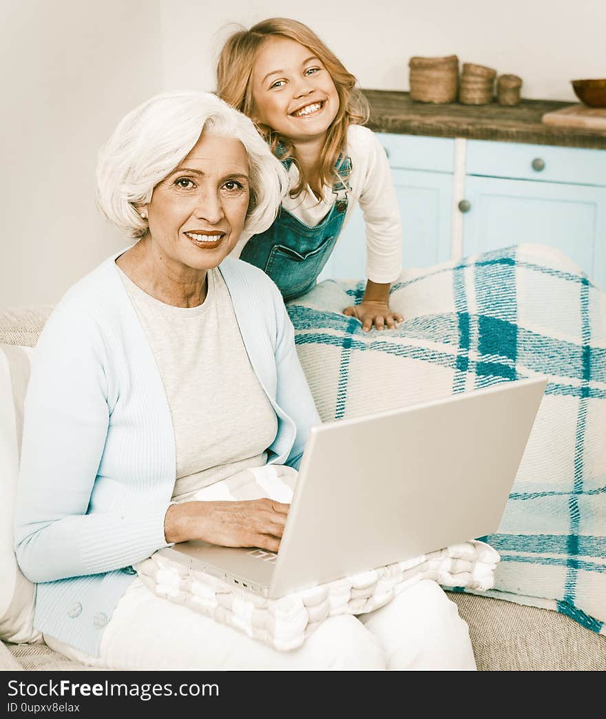 Smiling Granny And Her Granddaughter Using Wireless Internet At Home Mature Woman With Blond Hair Sitting On Sofa And Typing On Computer, Lady Working Or Messaging With Someone On Laptop Computer. Smiling Granny And Her Granddaughter Using Wireless Internet At Home Mature Woman With Blond Hair Sitting On Sofa And Typing On Computer, Lady Working Or Messaging With Someone On Laptop Computer