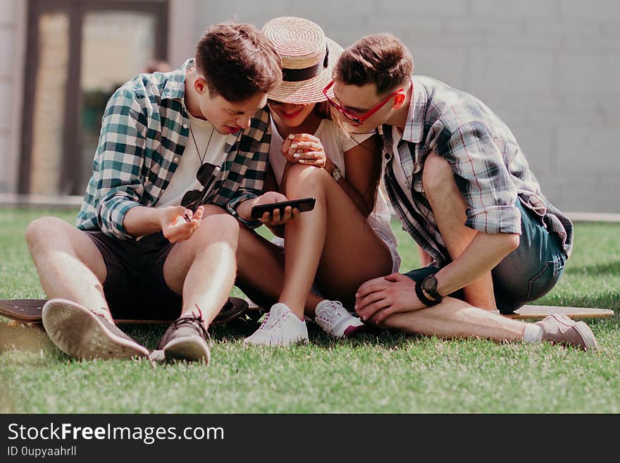 The Youngsters Leaned Together Over A Mobile Choosing The Best Photo Sitting On The Grass On A Summer Day. Stylish Young People In Casual. The Youngsters Leaned Together Over A Mobile Choosing The Best Photo Sitting On The Grass On A Summer Day. Stylish Young People In Casual.