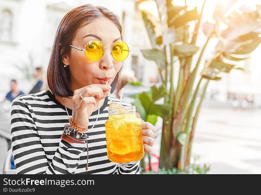 Female Hipster Student Drinks A Cool Lemonade Through A Straw In A Summer Outdoor Cafe