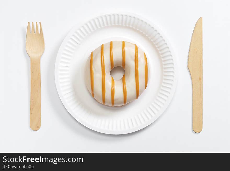 Glazed donut on a paper plate and wooden cutlery on a white background, top view. Glazed donut on a paper plate and wooden cutlery on a white background, top view