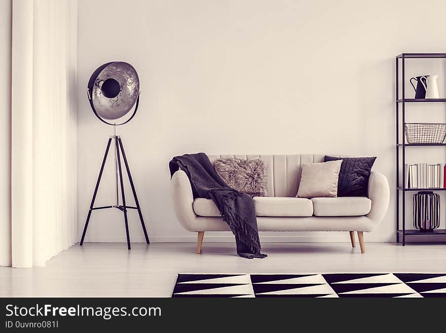 Black lamp next to beige sofa with blanket and pillows and shelf with books, copy space on empty white wall