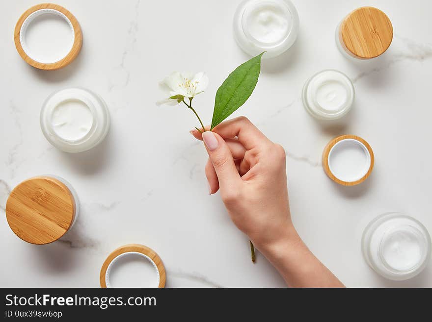 Cropped view of woman hand with jasmine flowers over jars with cosmetic cream on white surface