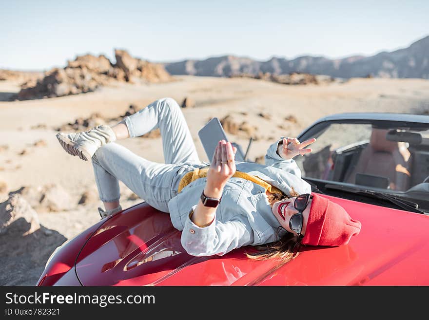 Lifestyle portrait of a young woman enjoying road trip on the desert valley, lying on the car hood and photographing on phone. Lifestyle portrait of a young woman enjoying road trip on the desert valley, lying on the car hood and photographing on phone