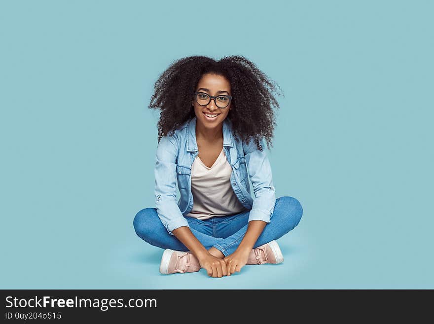 Young african girl wearing eyeglasses sitting on floor  on gray background leaning forward looking camera smiling joyful. Young african girl wearing eyeglasses sitting on floor  on gray background leaning forward looking camera smiling joyful