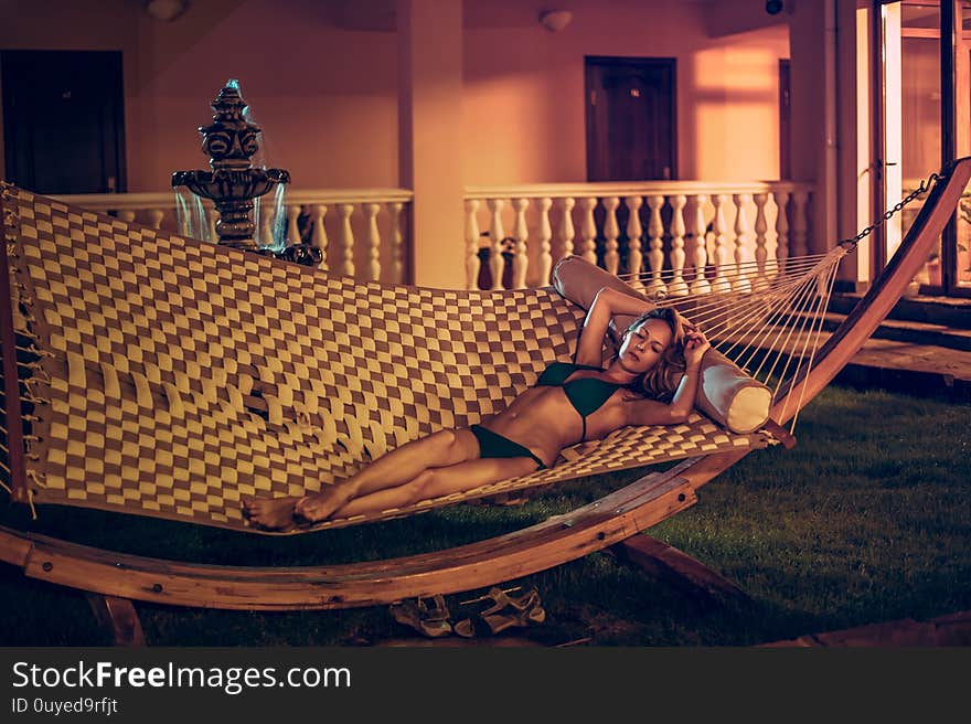 Slender beautiful young woman in a swimsuit is lying on a wicker hammock in the night yard of the garden of her house in summer. Slender beautiful young woman in a swimsuit is lying on a wicker hammock in the night yard of the garden of her house in summer