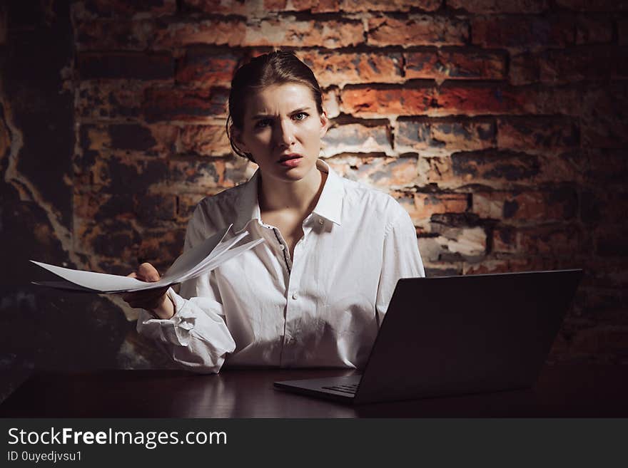 Displeased confused young female entrepreneur holding documents and looking inquiringly at camera while sitting at table with laptop against brick wall. Displeased confused young female entrepreneur holding documents and looking inquiringly at camera while sitting at table with laptop against brick wall