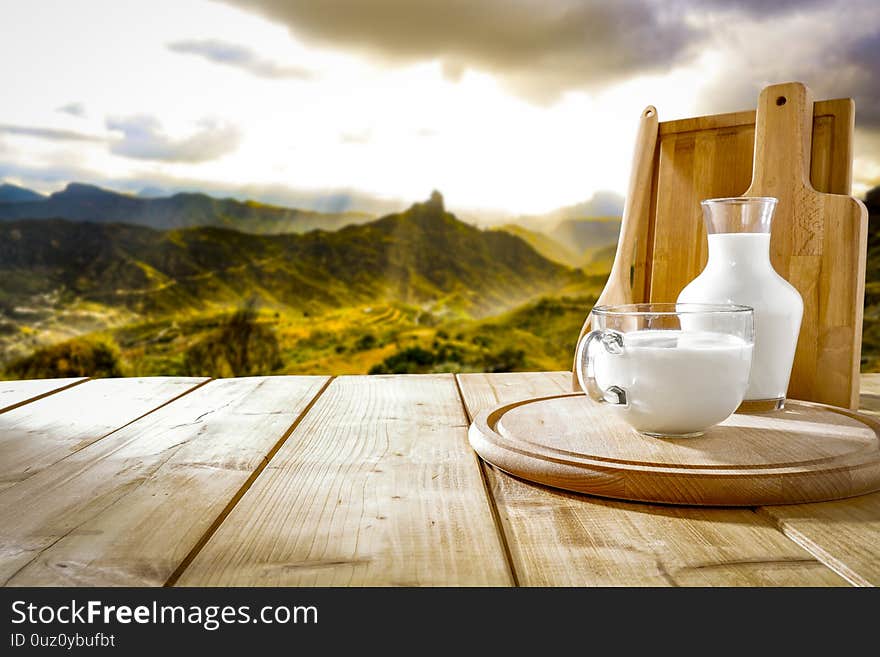 Wooden table top background with space for advertising product or decoration. White sky and mountains view in distance. Fresh milk in a glass and wooden cutting board on table top with mountains background and spring time. Wooden table top background with space for advertising product or decoration. White sky and mountains view in distance. Fresh milk in a glass and wooden cutting board on table top with mountains background and spring time.