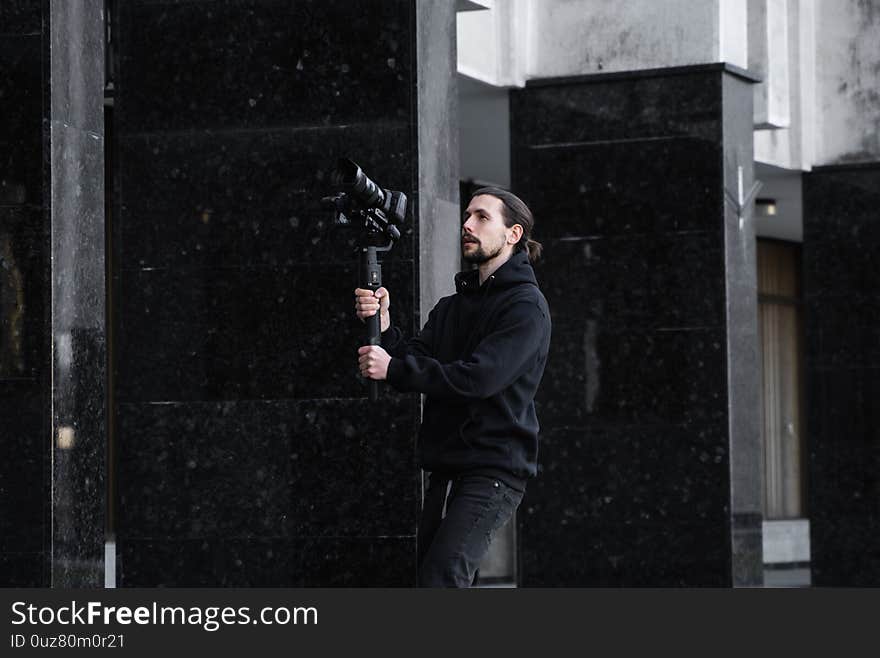 Young Professional videographer holding professional camera on 3-axis gimbal stabilizer. Pro equipment helps to make high quality video without shaking. Cameraman wearing black hoodie making a videos