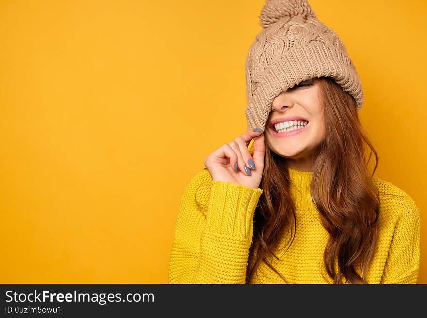 A brunette in a yellow sweater stands on a yellow background and smiles with white teeth, pulling the cap over her face. Horizontal photo