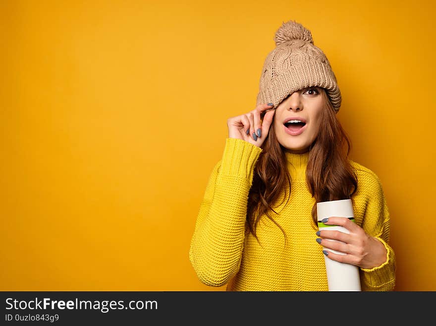 A brunette in a yellow sweater with a thermos is standing on a yellow background and pulling a hat over her face, her mouth open in surprise. Horizontal photo. A brunette in a yellow sweater with a thermos is standing on a yellow background and pulling a hat over her face, her mouth open in surprise. Horizontal photo