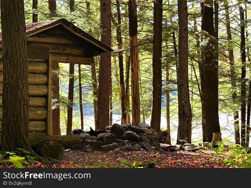 Small log cabin building in woodlands has campfire ring of boulders and rocks on autumn leaves with trees and lake in background.  Unpopulated, the structure sits ready for hikers and backpackers to settle in for an overnight to camp in the woods and mountains. Small log cabin building in woodlands has campfire ring of boulders and rocks on autumn leaves with trees and lake in background.  Unpopulated, the structure sits ready for hikers and backpackers to settle in for an overnight to camp in the woods and mountains.