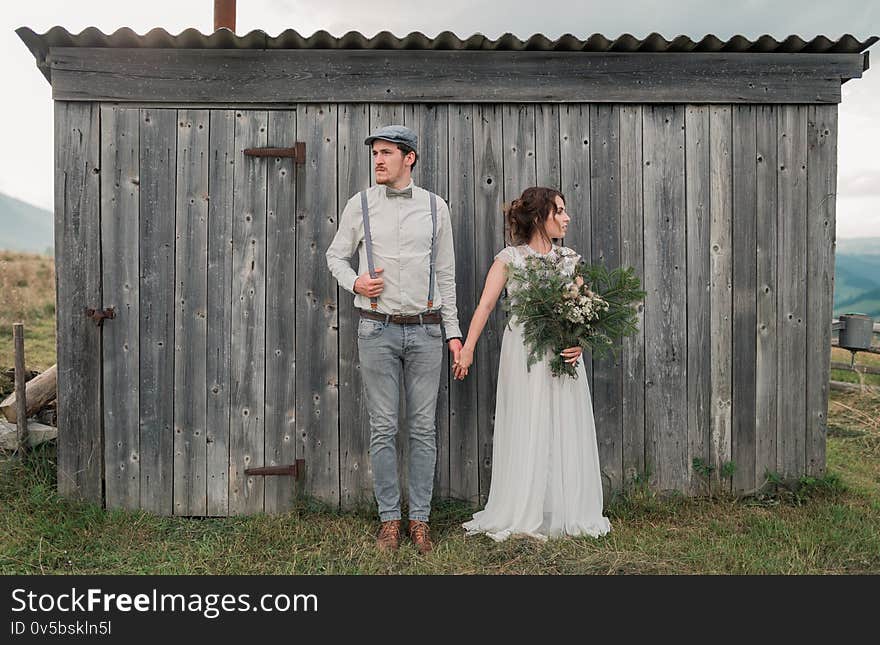 Style couple holding hands near gray wooden wall. In the fields at mountain sunset. Modern couple and ideas for the ceremony. Beautiful couple with mountains amazing view. Wedding in American style. Style couple holding hands near gray wooden wall. In the fields at mountain sunset. Modern couple and ideas for the ceremony. Beautiful couple with mountains amazing view. Wedding in American style.