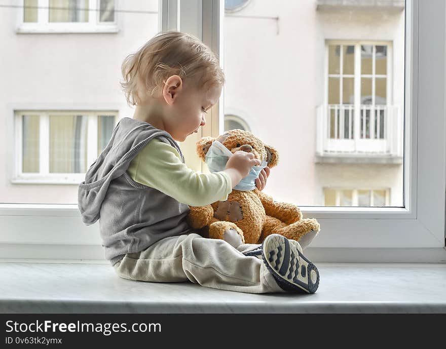 Child in home quarantine playing at the window with his sick teddy bear wearing a medical mask against viruses during coronavirus