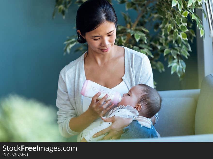 Happy young mother feeding her baby daughter with feeding bottle while sitting on sofa at home