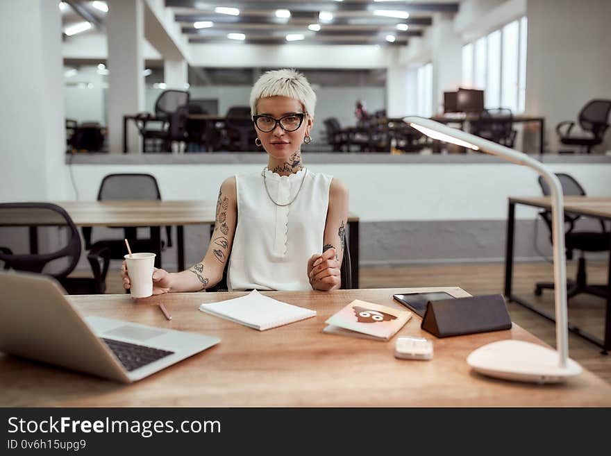 Young tattooed business woman in eyewear holding cup of coffee and looking at camera while working alone in the modern office. Business concept. Successful women. Coffee break. Young tattooed business woman in eyewear holding cup of coffee and looking at camera while working alone in the modern office. Business concept. Successful women. Coffee break